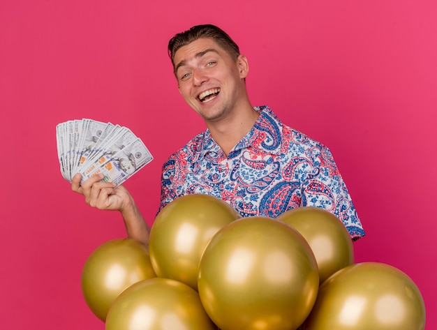 Smiling young party guy wearing colorful shirt standing behind balloons and holding cash isolated on pink