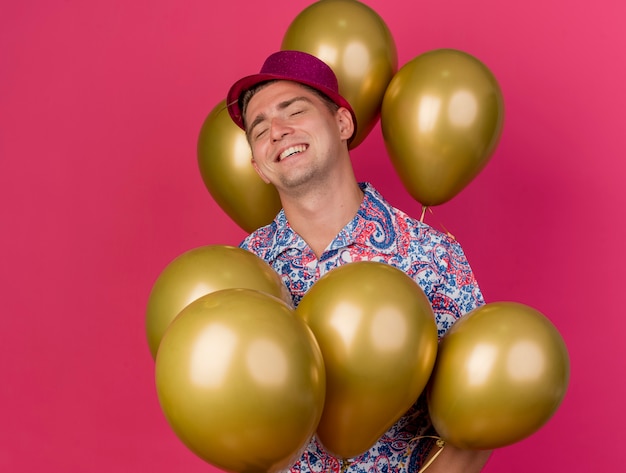 Smiling young party guy with closed eyes wearing pink hat standing among balloons isolated on pink