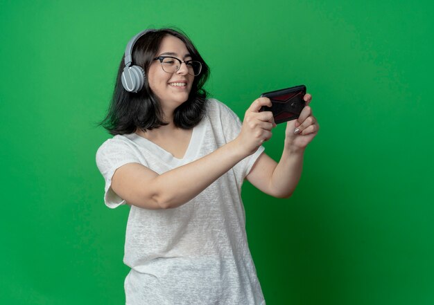 Smiling young pretty caucasian girl wearing glasses and headphones holding and looking at mobile phone isolated on green background with copy space