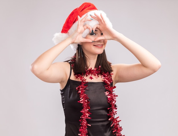 Free photo smiling young pretty caucasian girl wearing santa hat and tinsel garland around neck looking at camera doing heart sign in front of eye isolated on white background