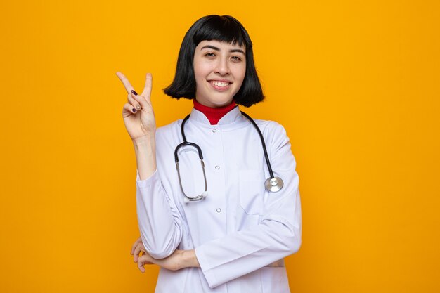 Smiling young pretty caucasian woman in doctor uniform with stethoscope gesturing victory sign