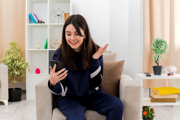 Smiling young pretty caucasian woman sitting on armchair in designed living room holding and looking at mobile phone and showing empty hand