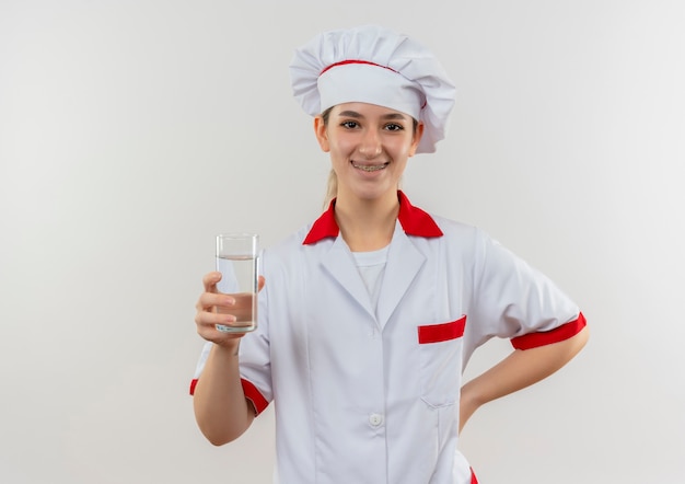 Smiling young pretty cook in chef uniform with dental braces holding glass of water and putting hand on waist isolated on white space