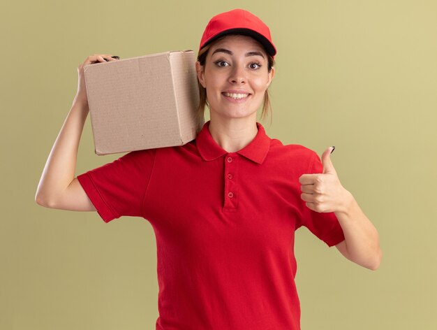 Smiling young pretty delivery girl in uniform thumbs up and holds cardbox on shoulder on olive green