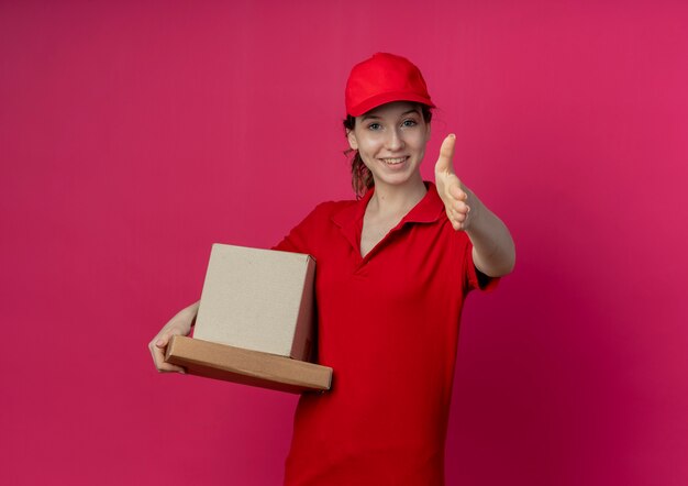 Smiling young pretty delivery girl wearing red uniform and cap holding pizza package and carton box stretching out hand towards camera gesturing hi isolated on crimson background with copy space