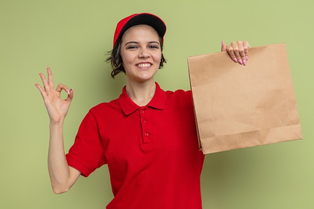 Smiling young pretty delivery woman holding paper food packaging and gesturing ok sign