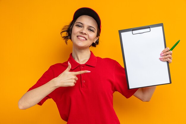 Smiling young pretty delivery woman holding and pointing at clipboard 