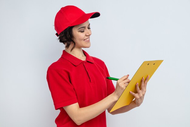 Smiling young pretty delivery woman holding and writing on clipboard 