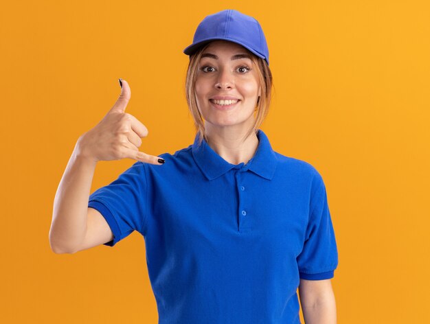 Smiling young pretty delivery woman in uniform doing hang loose gesture isolated on orange wall