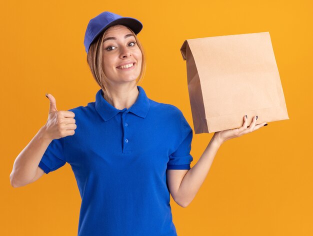 Smiling young pretty delivery woman in uniform thumbs up and holds paper package isolated on orange wall