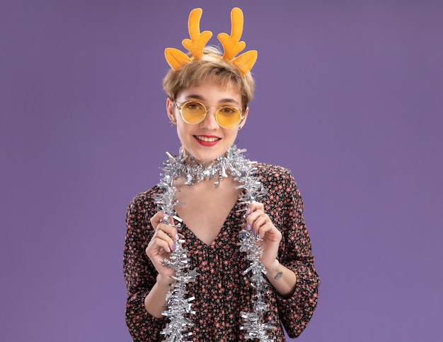 Free photo smiling young pretty girl wearing reindeer antlers headband and tinsel garland around neck with glasses  grabbing tinsel garland isolated on purple wall with copy space