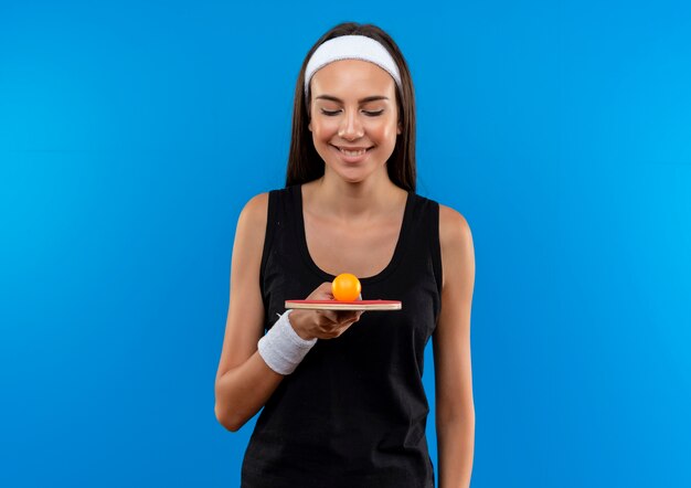 Smiling young pretty sporty girl wearing headband and wristband holding and looking at ping pong racket with ball on blue space