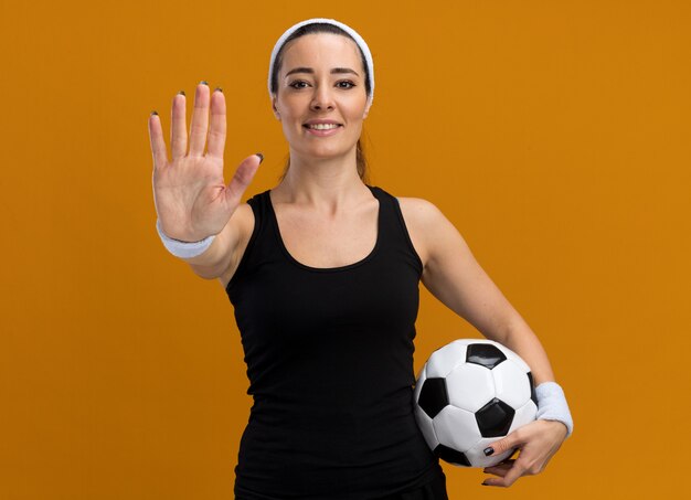 Smiling young pretty sporty girl wearing headband and wristbands holding soccer ball doing stop gesture 