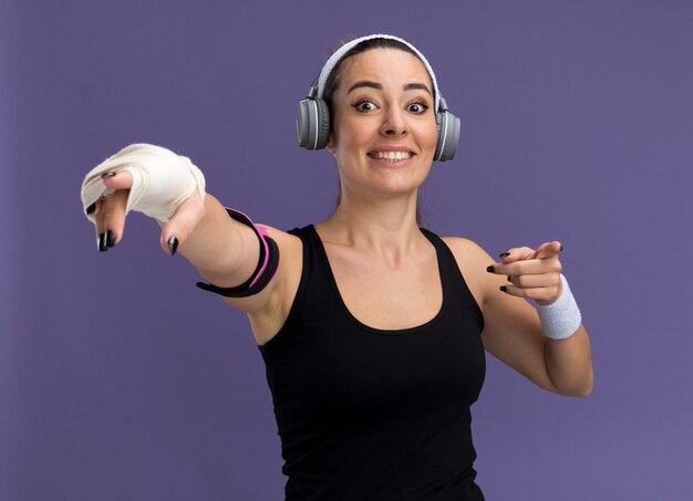 Smiling young pretty sporty woman wearing headband wristbands headphones and phone armband with injured wrist wrapped with bandage looking and pointing at front isolated on purple wall