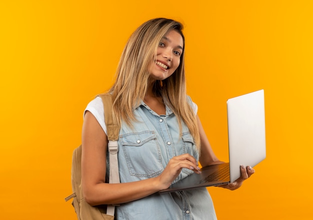 Smiling young pretty student girl wearing back bag holding and using laptop isolated on orange wall