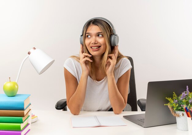 Smiling young pretty student girl wearing headphones sitting at desk with school tools listening to music looking at side with fingers on headphones isolated on white wall