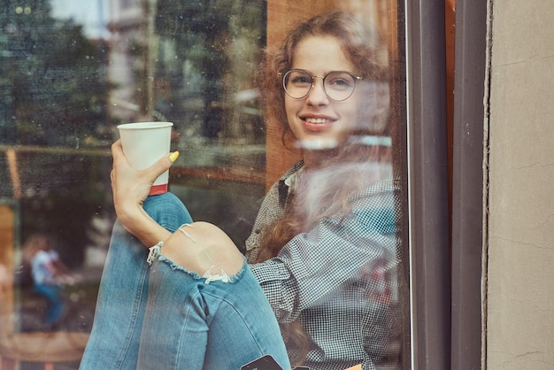 Smiling young redhead curly girl wearing casual clothes and glasses sitting on a window sill with a takeaway coffee.