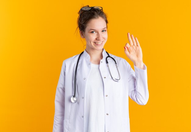 Smiling young slavic girl in doctor uniform with stethoscope gesturing ok sign isolated on orange wall with copy space