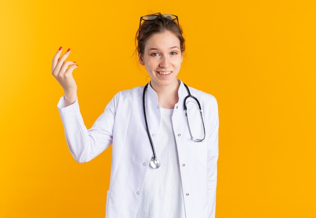 Smiling young slavic girl in doctor uniform with stethoscope pretending to hold something isolated on orange wall with copy space