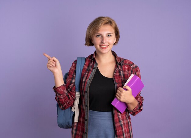 Smiling young slavic student girl wearing backpack holds book and notebook points at side 