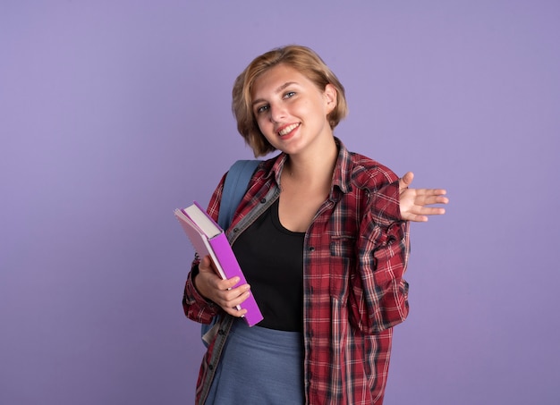 Smiling young slavic student girl wearing backpack keeps hand open holds book and notebook 