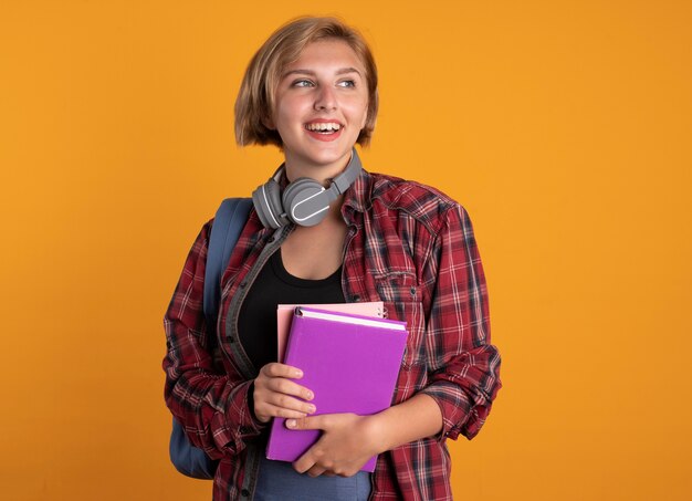 Smiling young slavic student girl with headphones around neck wearing backpack holds book and notebook looking at side 