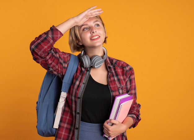 Smiling young slavic student girl with headphones wearing backpack keeps palm at forehead holds book and notebook 