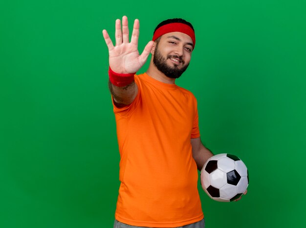 Smiling young sporty man wearing headband and wristband holding ball showing five isolated on green background with copy space