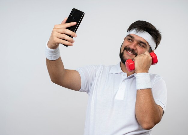 Smiling young sporty man wearing headband and wristband holding dumbbell take a selfie isolated on white wall