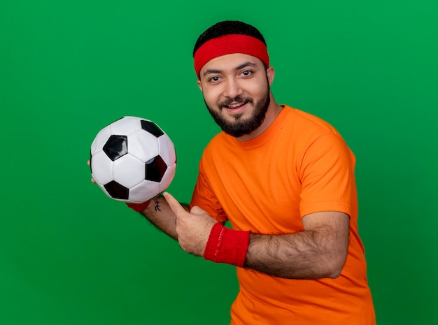 Smiling young sporty man wearing headband and wristband holding and points at ball isolated on green background