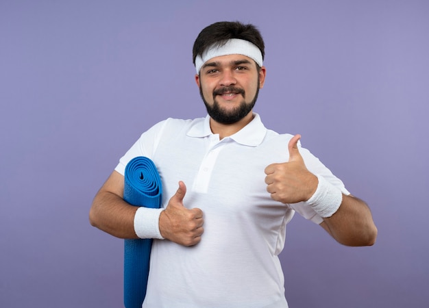 Smiling young sporty man wearing headband and wristband holding yoga mat showing thumbs up