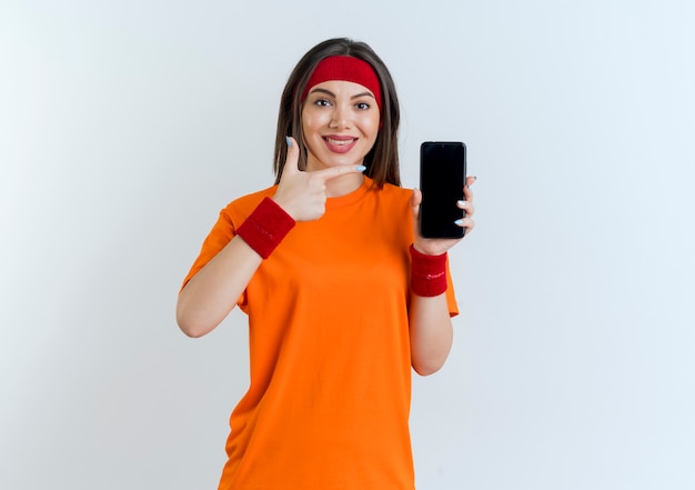 Smiling young sporty woman wearing headband and wristbands showing mobile phone pointing at it looking isolated