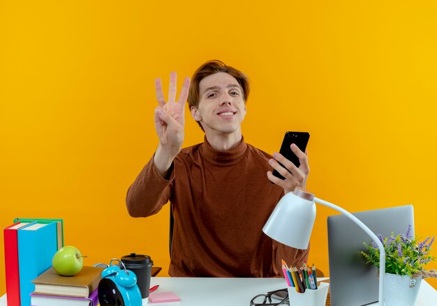 Smiling young student boy sitting at desk with school tools holding phone and showing three isolated on yellow wall