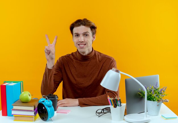 Smiling young student boy sitting at desk with school tools peace gesture on yellow