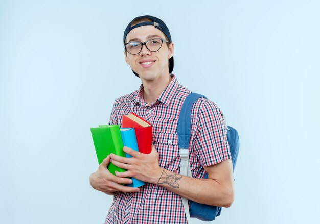 Smiling young student boy wearing back bag and glasses and cap holding books on white