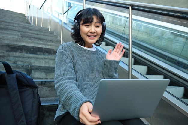 Smiling young student wears headphones sits on street stairs and waves hand at her laptop connects t