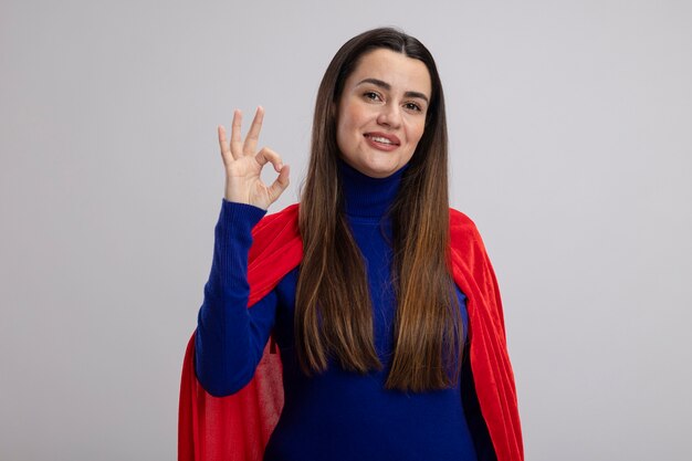 Smiling young superhero girl showing okay gesture isolated on white