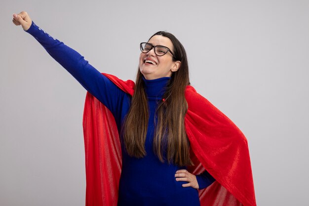 Smiling young superhero girl wearing glasses raising fist isolated on white