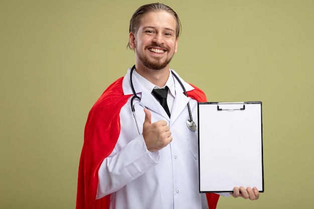 Free photo smiling young superhero guy wearing stethoscope with medical robe holding clipboard showing thumb up isolated on olive green background