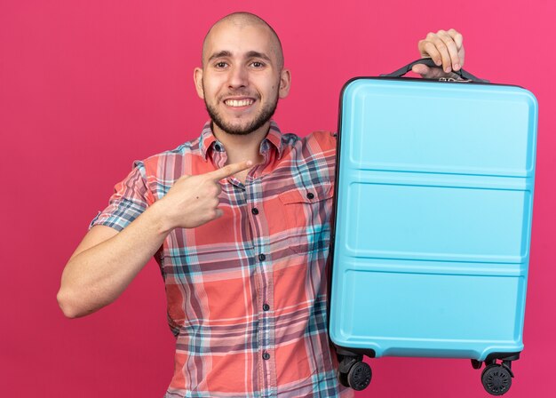 Free Photo smiling young traveler man holding and pointing at suitcase isolated on pink wall with copy space