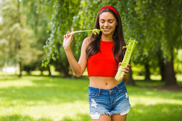 Smiling young woman eating fresh celery in the park