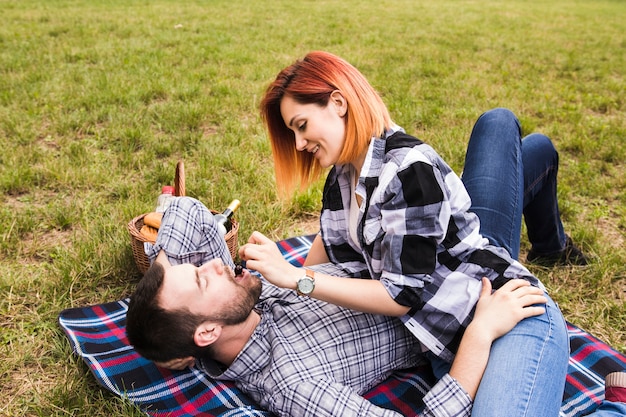 Free photo smiling young woman feeding cherry to her boyfriend lying on blanket over green grass