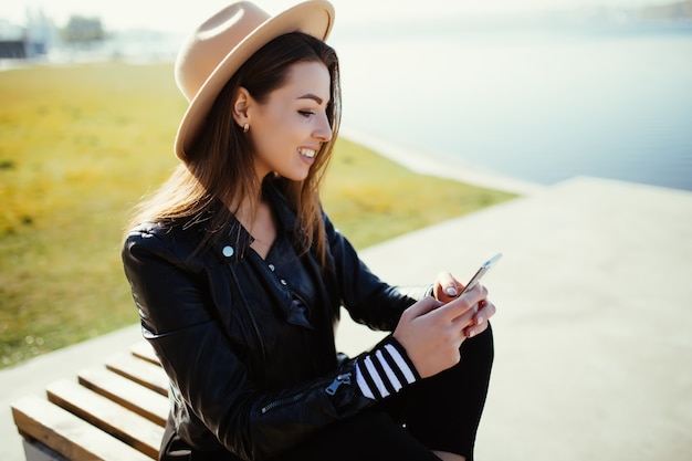 Smiling young woman girl sitting in the park near the city lake in cold sunny summer day dressed up in black clothes