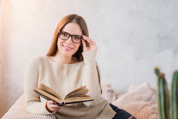 Free photo smiling young woman holding book in hand sitting against wall