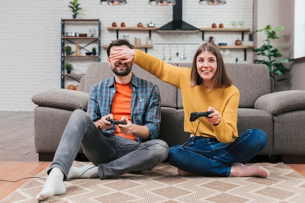 Smiling young woman holding joystick covering her husband's eyes while playing the video game