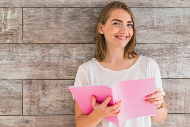 Smiling young woman holding pink book against wooden wall