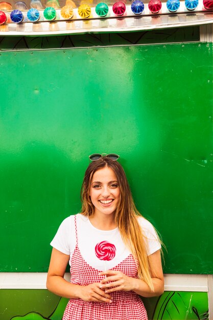 Smiling young woman holding red lollipop standing in front of green wall