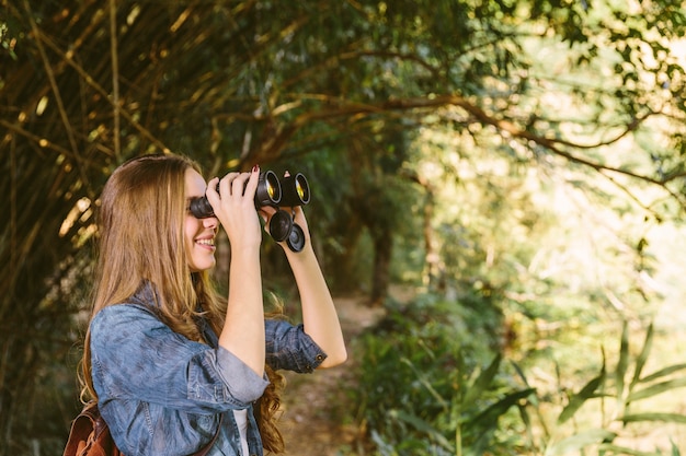 Free photo smiling young woman looking through binoculars in forest