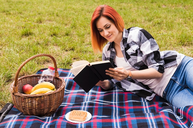 Free photo smiling young woman lying on blanket reading book at picnic