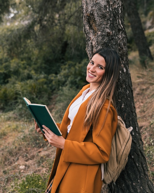 Smiling young woman posing under the tree with book in her hand